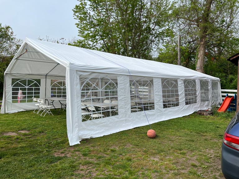 Large white event tent with clear windows set up on a grassy area with trees, vehicles, and outdoor furniture visible