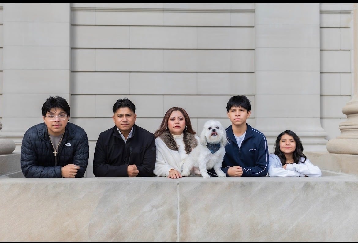 Five people and a white fluffy dog lined up against a white columned building background