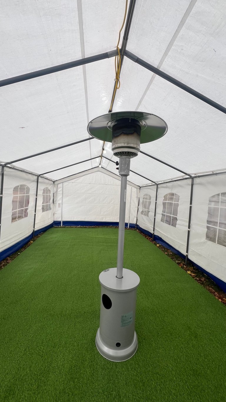 Patio heater with green shade mounted under a clear-top tent pavilion with white walls and green artificial turf flooring