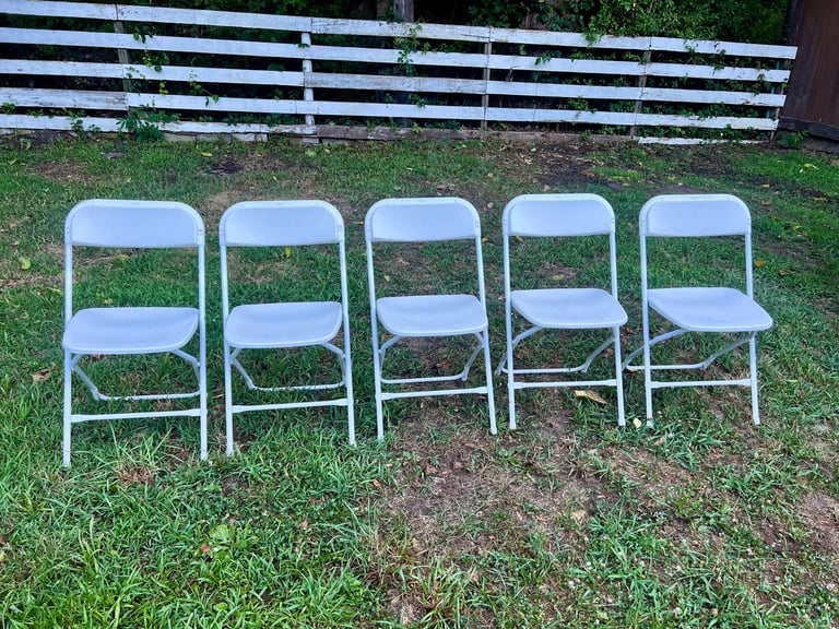 Five white folding chairs lined up on grass with a striped fence in the background