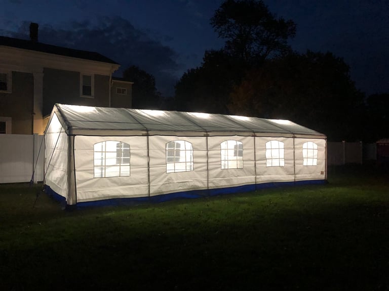 Illuminated white party tent with blue base set up on grass at dusk near a residential building