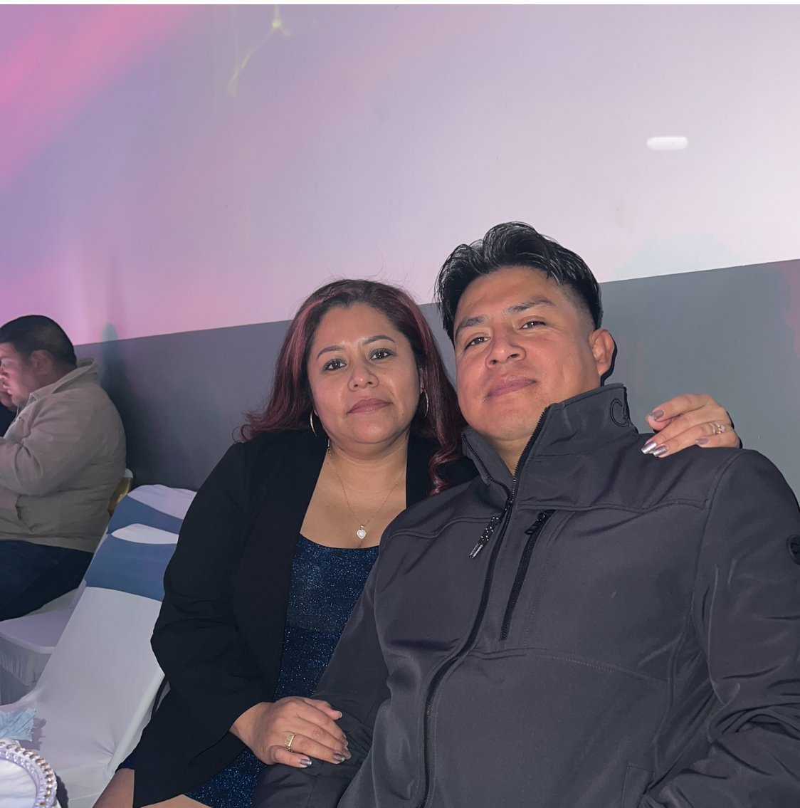 A couple posing together indoors with a pink and purple gradient background. The man wears a black jacket and the woman wears a black blazer over a blue top.