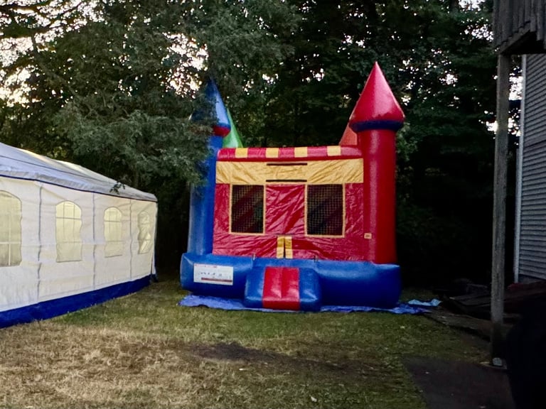 Red and blue inflatable bounce castle with red turrets in a backyard surrounded by trees and residential structures